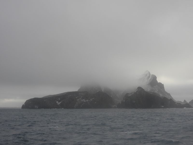 The image depicts a rugged, snow-covered mountainous island partially obscured by fog and clouds. Th…